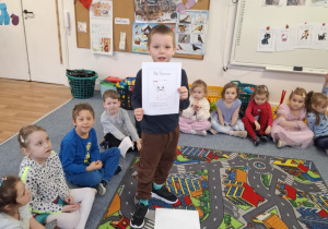 Chłopiec stoi na dywanie i pokazuje dzieciom obrazek z kotem z bajki