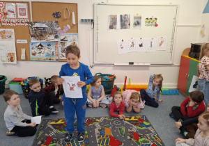 Chłopiec stoi na dywanie i pokazuje dzieciom obrazek z kotem z bajki