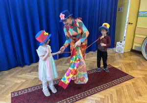 Pani w kolorowym stroju i dwójka dzieci prezentują sztuczkę iluzjonistyczną