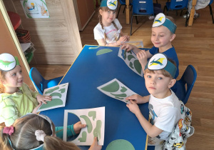 Grupa dzieci siedzi przy stoliku i wypycha z kartonu postać dinozaura