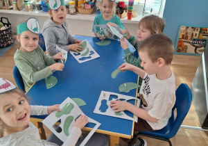 Grupa dzieci siedzi przy stoliku i wypycha z kartonu postać dinozaura