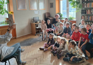 Dzieci siedzą na ławkach i słuchają pani bibliotekarki czytającej książkę