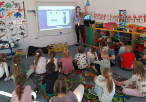 Dzieci siedzą na dywanie i słuchają pani prowadzącej warsztaty