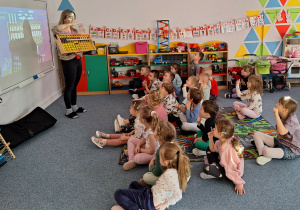 Dzieci siedzą na dywanie i słuchają pani prowadzącej warsztaty