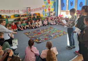 Dzieci siedzą na dywanie wraz z wychowawczynią i słuchają czytającego chłopca