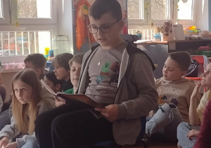 Uczeń siedzi na krześle i czyta wiersz przedszkolakom