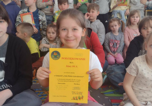 Uczennica trzyma dyplom i pozuje do zdjęcia z przedszkolakami i uczniami