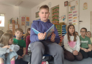Uczeń siedzi na krześle i czyta wiersz przedszkolakom
