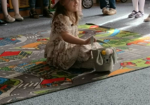 Dziewczynka"Pani wiosna" siedzi na dywanie a dzieci stoją na dywanie i śpiewają piosenkę