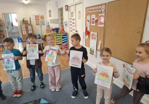 Grupa dzieci stoi na dywanie i pokazuje laurki dla Ani