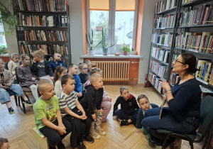Dzieci siedzą na ławeczkach i słuchają czytanej przez p.bibliotekarkę książkę