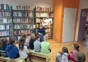 Dzieci siedzą na ławeczkach i słuchają czytanej przez p.bibliotekarkę książkę