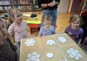Grupa dzieci stoi przy stolikach i słucha instrukcji do wykonania tęczowego kwiatka