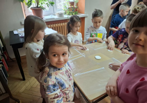 Grupa dzieci stoi przy stolikach i słucha instrukcji do wykonania tęczowego kwiatka