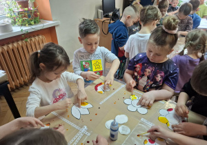 Grupa dzieci stoi przy stolikach i maluje tęczowe kwiaty