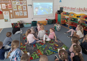 Dzieci siedzą na dywanie i oglądają film edukacyjny na tablicy multimedialnej
