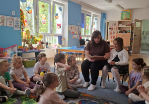 Dzieci siedzą na dywanie i słuchają czytającej mamy i siostry Ani