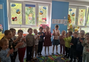 Grupa dzieci stoi na dywanie i śpiewa Krzysiowi sto lat