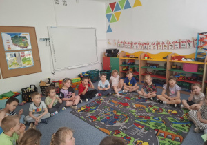 Dzieci siedzą na dywanie i słuchają opowiadania z książki, które czyta babcia Wojtka