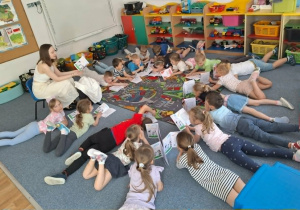 Dzieci leżą na dywanie z książeczkami i słuchają opowiadania pani prowadzącej