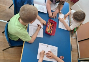 Grupa dzieci siedzi przy stolikach i koloruje obrazki w książeczce