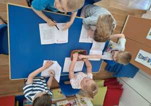 Grupa dzieci siedzi przy stolikach i koloruje obrazki w książeczce