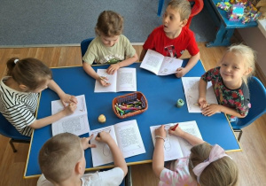 Grupa dzieci siedzi przy stolikach i koloruje obrazki w książeczce