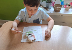 Chłopiec siedzi przy stole i maluje farbami ceramiczny kwiatek