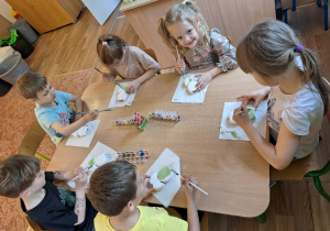 Grupa dzieci siedzi przy stole i maluje farbami ceramiczny kwiatek