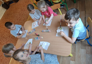 Grupa dzieci siedzi przy stole i maluje farbami ceramiczny kwiatek