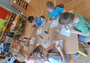 Grupa dzieci siedzi przy stole i maluje farbami ceramiczny kwiatek