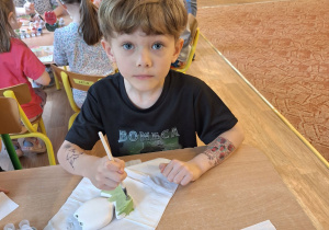 Chłopiec siedzi przy stole i maluje farbami ceramiczny kwiatek