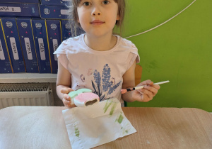 Dziewczynka siedzi przy stole i maluje farbami ceramiczny kwiatek