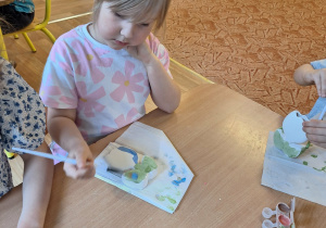 Dziewczynka siedzi przy stole i maluje farbami ceramiczny kwiatek
