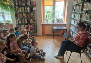 Dzieci siedzą na ławeczkach i słuchają jak pani bibliotekarka czyta im książkę