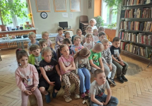 Dzieci siedzą na ławeczkach i słuchają jak pani bibliotekarka czyta im książkę