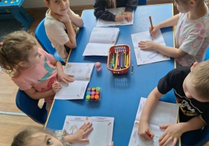 Grupa dzieci siedzi przy stoliku i koloruje książeczkę
