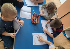 Grupa dzieci siedzi przy stoliku i koloruje książeczkę