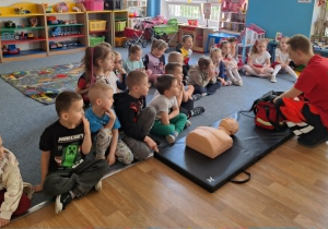 Dzieci siedzą na dywanie i słuchają ratownika medycznego który opowiada im o swojej pracy