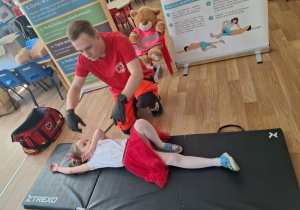 Ratownik medyczny pokazuje na dziewczynce jak wyląda ułożenie ciała w bezpiecznej pozycji