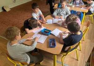Dzieci siedzą przy stolikach i rysują w książkach
