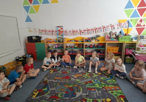 Dzieci siedzą na dywanie i wspólnie bawią się z panią prowadzącą