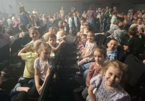 Grupa dzieci siedzi w teatrze na krzesłach i czeka na przedstawienie