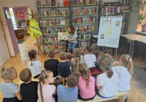 Dzieci siedzą na ławkach i słuchają p.bibliotekarki czytającej im książkę