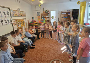 Chłopcy siedzą na krzesłach, a dziewczynki stojąc na dywanie śpiewają im sto lat