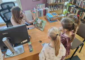 Dziewczyki stoją i słuchają pani bibliotekarki, w jaki sposób można wypożyczyć książkę