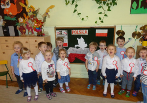 Dzieci śpiewają hymn przy tablicy z symbolami narodowymi.