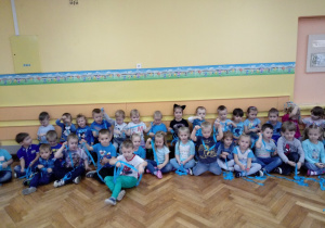 Grupa dzieci siedzi na podłodze, trzymając w dłoniach niebieskie wstążki