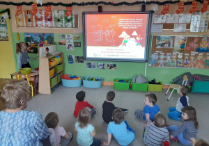 Dzieci siedzą na dywanie i oglądają prezentację multimedialną