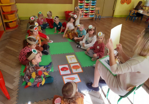 Dzieci siedzą w kole i oglądają ilustracje warzyw pokazywane przez nauczycielkę